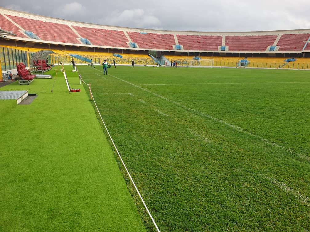 Bir futbol stadyumunun yeşil çim alanı, kenarlarda yedek koltuklar ve oyuncuların çalıştığı bir sahne.